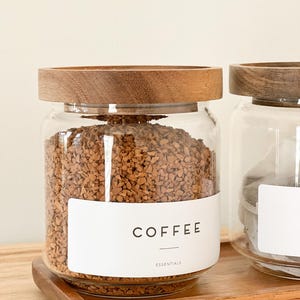 May include: A glass jar with a wooden lid filled with coffee grounds. The jar has a white label that says "COFFEE" and "ESSENTIALS".