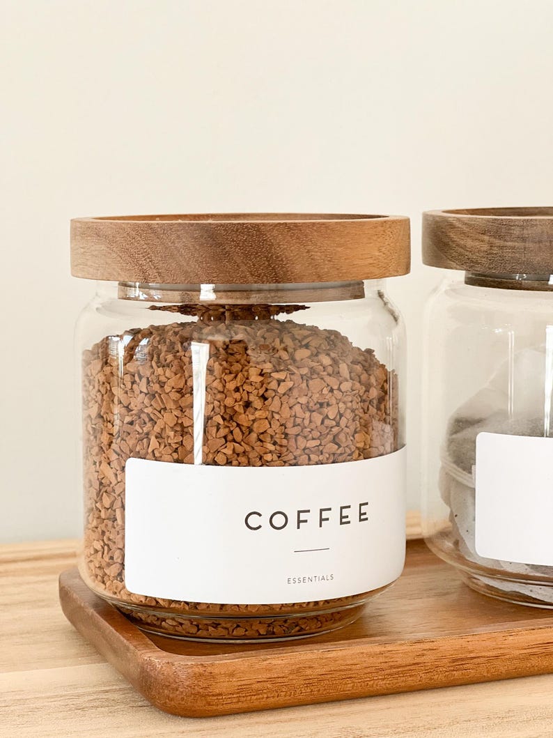 May include: A glass jar with a wooden lid filled with coffee grounds. The jar has a white label that says "COFFEE" and "ESSENTIALS".