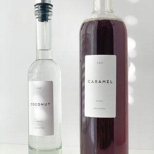 Puede incluir: Dos botellas de vidrio con etiquetas. La botella de la izquierda es transparente y contiene un l&iacute;quido blanco. La etiqueta dice "SYRUP COCONUT 100% NATURAL". La botella de la derecha es marr&oacute;n y contiene un l&iacute;quido oscuro. La etiqueta dice "SYRUP CARAMEL 100% NATURAL".