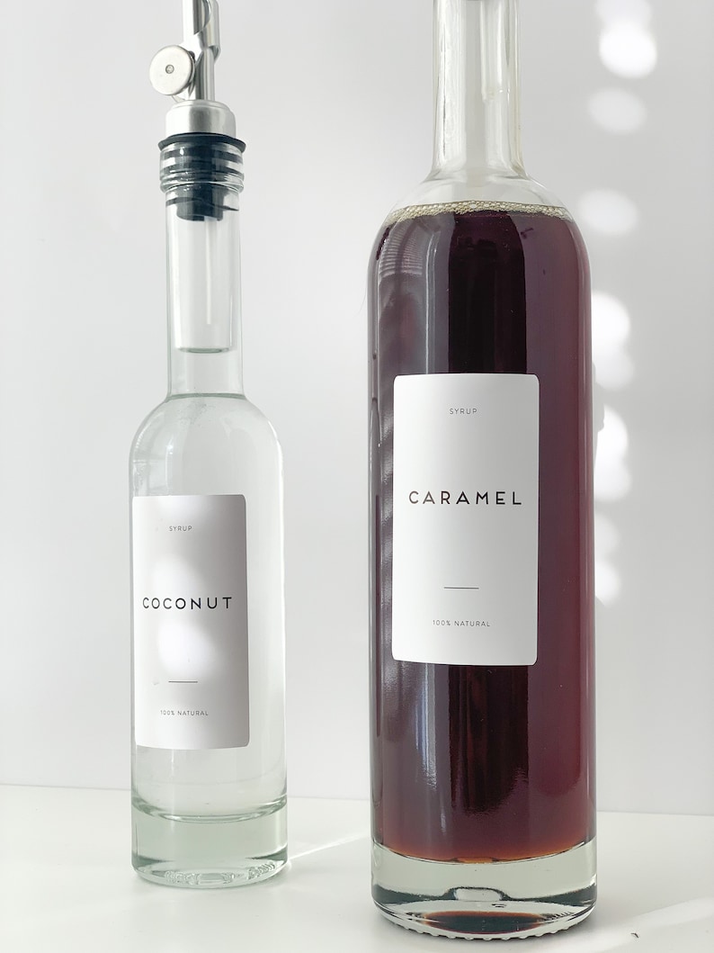 Puede incluir: Dos botellas de vidrio con etiquetas. La botella de la izquierda es transparente y contiene un l&iacute;quido blanco. La etiqueta dice "SYRUP COCONUT 100% NATURAL". La botella de la derecha es marr&oacute;n y contiene un l&iacute;quido oscuro. La etiqueta dice "SYRUP CARAMEL 100% NATURAL".