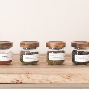 Könnte beinhalten: Vier Glasbehälter mit Holzdeckeln und Etiketten. Die Behälter enthalten getrocknete Kräuter und Gewürze, darunter Chilipulver, Petersilie, Oregano und schwarzer Pfeffer.