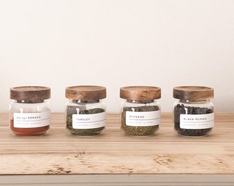Peut inclure: Quatre bocaux en verre avec des couvercles en bois et des étiquettes. Les bocaux contiennent des herbes et des épices séchées, dont du piment en poudre, du persil, de l'origan et du poivre noir.