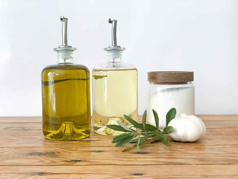 May include: Two glass bottles with pour spouts filled with olive oil and cooking oil, a glass jar with a wooden lid filled with salt, a garlic clove, and a sprig of green olive leaves on a wooden surface.