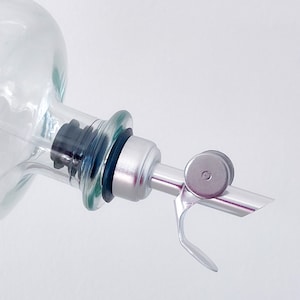 K&ouml;nnte beinhalten: Eine klare Glasflasche mit einem silbernen Ausgie&szlig;er aus Metall und einem schwarzen Gummistopfen. Der Ausgie&szlig;er hat einen kleinen, runden, silbernen Knopf.