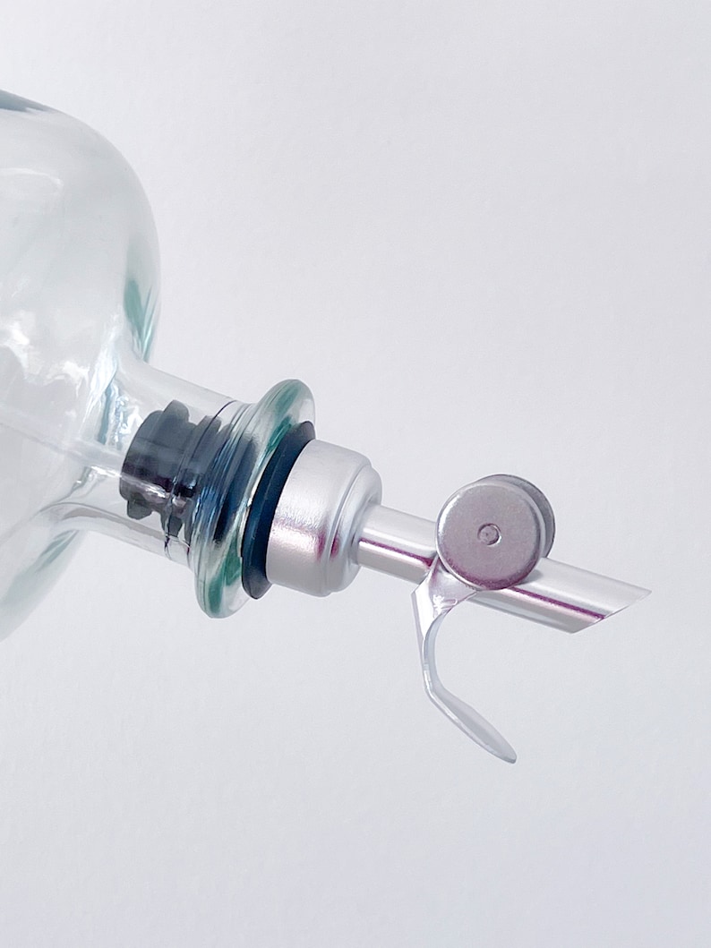 K&ouml;nnte beinhalten: Eine klare Glasflasche mit einem silbernen Ausgie&szlig;er aus Metall und einem schwarzen Gummistopfen. Der Ausgie&szlig;er hat einen kleinen, runden, silbernen Knopf.
