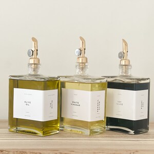 May include: Three glass bottles with gold spouts and labels. The bottles contain olive oil, white vinegar, and soy sauce. The labels are white with black text.