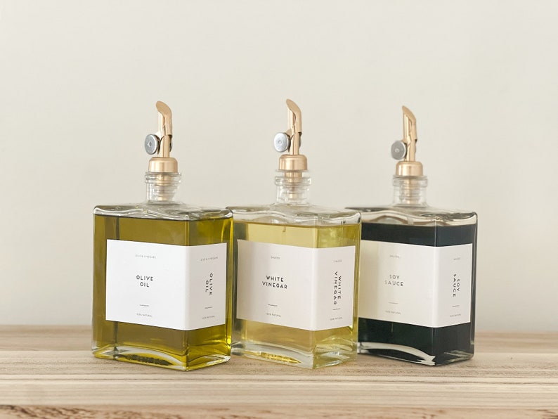 May include: Three glass bottles with gold spouts and labels. The bottles contain olive oil, white vinegar, and soy sauce. The labels are white with black text.