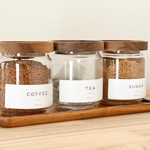 May include: Three glass jars with wooden lids, labeled "Coffee", "Tea", and "Sugar". The jars are filled with coffee, tea bags, and sugar, respectively. The jars are sitting on a wooden tray.