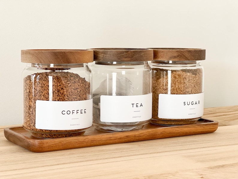 May include: Three glass jars with wooden lids, labeled "Coffee", "Tea", and "Sugar". The jars are filled with coffee, tea bags, and sugar, respectively. The jars are sitting on a wooden tray.
