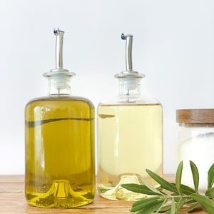 May include: Two glass bottles with pour spouts filled with olive oil and cooking oil, sitting on a wooden surface with a sprig of green leaves.
