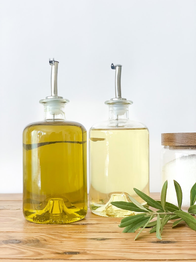 May include: Two glass bottles with pour spouts filled with olive oil and cooking oil, sitting on a wooden surface with a sprig of green leaves.
