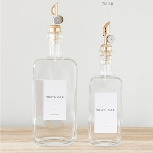 May include: Two clear glass bottles with gold pump tops and white labels that say "MOUTHWASH YOUR NATURAL" and "MOUTHWASH YOUR NATURAL". The bottles are on a light brown wooden surface. The larger bottle is labelled 500ml and the smaller bottle is labelled 200ml.