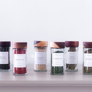 May include: Six glass spice jars with wooden lids, each filled with a different spice or herb. The labels on the jars read "Black Pepper", "Paprika", "Sesame Seeds", "Parsley", "Bay Leaves", and "Oregano".