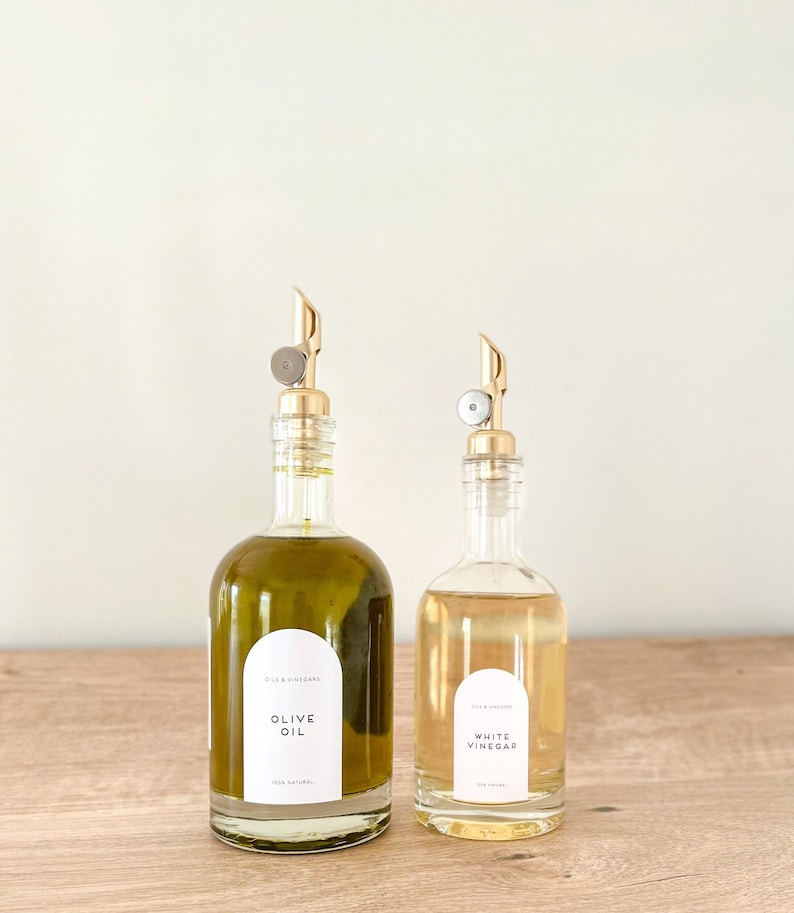 K&ouml;nnte beinhalten: Zwei Glasflaschen mit goldenen Ausgie&szlig;ern und Etiketten. Die Flasche links ist mit Oliven&ouml;l gef&uuml;llt und mit "Oliven&ouml;l" beschriftet. Die Flasche rechts ist mit wei&szlig;em Essig gef&uuml;llt und mit "Wei&szlig;er Essig" beschriftet. Beide Flaschen stehen auf einer Holzoberfl&auml;che.