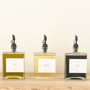 May include: Three glass bottles with black pump tops and white labels. The bottles contain olive oil, white vinegar, and soy sauce. The bottles are sitting on a wooden surface.