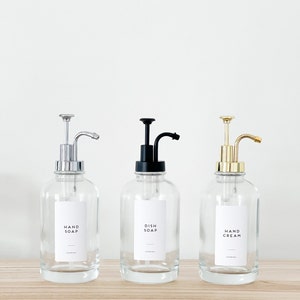 May include: Three glass bottles with pumps and labels. The bottles are filled with liquid. The labels read "Hand Soap", "Dish Soap", and "Hand Cream".