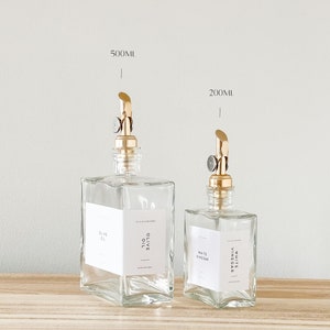 May include: Two clear glass bottles with gold pump tops and labels. The larger bottle is labeled "Olive Oil" and the smaller bottle is labeled "White Vinegar". The bottles are sitting on a wooden surface.