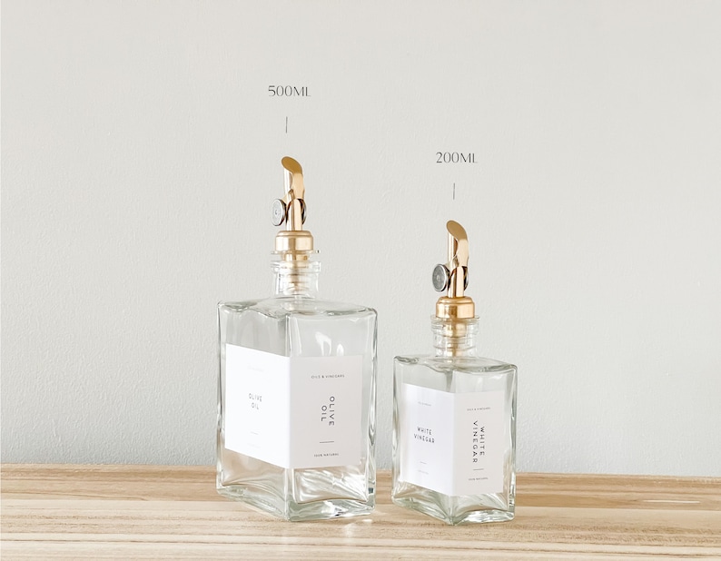May include: Two clear glass bottles with gold pump tops and labels. The larger bottle is labeled "Olive Oil" and the smaller bottle is labeled "White Vinegar". The bottles are sitting on a wooden surface.