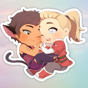 Op de afbeelding: Een sticker met twee stripfiguren in een liefdevolle omhelzing. De ene heeft bruin haar en kattenoren, met een paarse tanktop. De andere heeft blond haar, met een rood topje en grijze broek. De sticker heeft een pastelkleurige achtergrond.