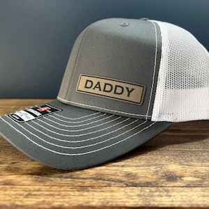 May include: A gray and white trucker hat with a brown leather patch that says "DADDY" on it.