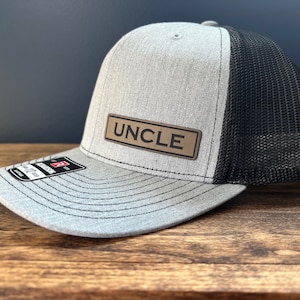 May include: A gray and black trucker hat with a brown leather patch that says "UNCLE" on it.