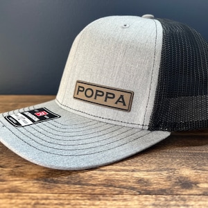 May include: A gray and black trucker hat with a brown leather patch that says "POPPA" on it.