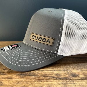 May include: A gray and white trucker hat with a brown leather patch that says "BUBBA" on it.