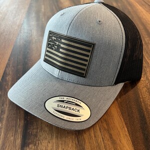 May include: A gray and black trucker hat with a black and white American flag patch featuring a silhouette of guns. The hat has a black mesh back and a gold label that reads "The Authentic Yupoong Snapback Yupoong Since 1974".