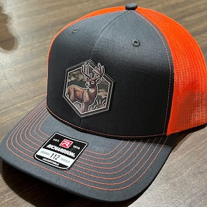 May include: A gray and orange trucker hat with a deer patch. The hat features a black front panel with a deer emblem, an orange mesh back, and a gray brim with orange stitching. The Richardson brand label is visible.