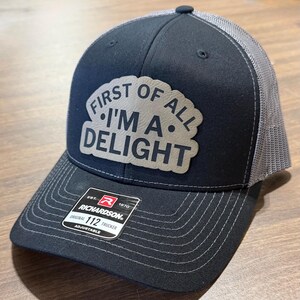 May include: Black and gray trucker hat with the text "FIRST OF ALL I'M A DELIGHT" on a patch. The hat has a curved brim and a mesh back. The Richardson brand tag is visible.