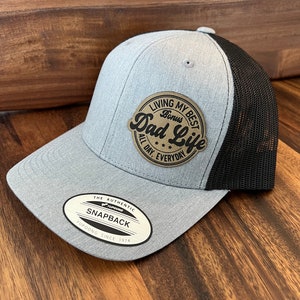 May include: Gray and black mesh trucker hat with a brown leather patch that says "Living My Best Dad Life Bonus All Day, Everyday". The hat has a black mesh back and a gray front with a white label that says "The Authentic YUPOONG Snapback YUPOONG Since 1974".