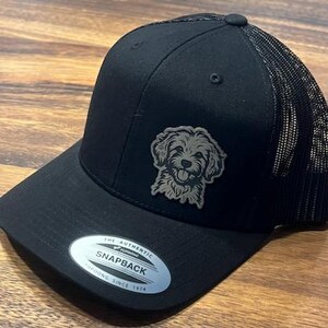 May include: Black trucker hat with a black mesh back and a gray embroidered image of a dog. The hat has a black brim and a white sticker on the underside of the brim that says "The Authentic Yupoong Snapback Founded Since 1974."