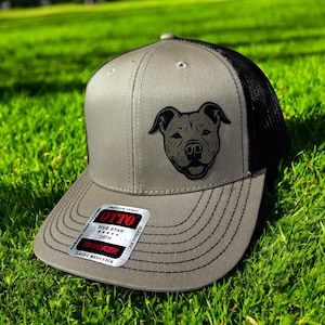May include: A gray and black trucker hat with a black embroidered image of a pit bull dog's face. The hat has a mesh back and a label that reads "Otto Five Star Trucker Classic Mesh Back".