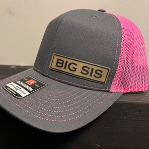 May include: Gray and pink trucker hat with the text "BIG SIS" on a leather patch. The hat has a mesh back and a curved brim. The stitching is pink, and the hat is adjustable.