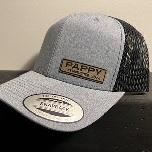 May include: A gray and black Snapback hat with the text "PAPPY ESTABLISHED 2024" on a brown patch. The hat has a curved brim and a mesh back. The tag reads "THE AUTHENTIC YUPOONG SNAPBACK YUPOONG SINCE 1974".