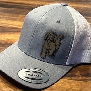 May include: A gray and white trucker hat with a brown felt patch of a doodle dog on the front. The hat has a black snap closure and a white mesh back.