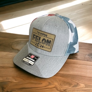 May include: A gray and blue trucker hat with a gold patch that says "I'm voting FELON -2024-". The hat has a red, white, and blue American flag design on the back.