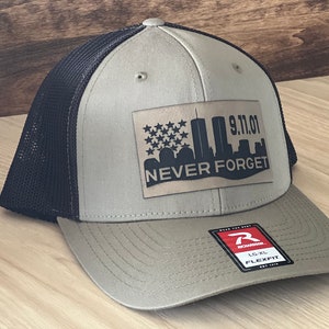 May include: A khaki and black trucker hat with a brown leather patch that says "9.11.01 NEVER FORGET" with a silhouette of the Twin Towers and the American flag. The hat is a Richardson brand with a Flexfit logo.