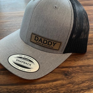 May include: Gray and black mesh baseball cap with a brown leather patch that says "Daddy Aiden & Lochlan". The cap has a black mesh back and a gray front with a white logo that says "The Authentic Yupoong Snapback Yupoong Since 1974".