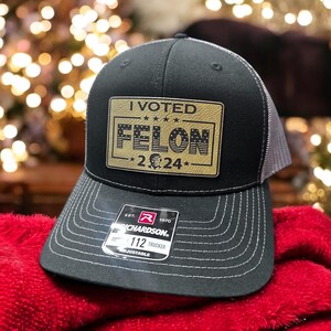 May include: A black and gray trucker hat with a gold patch that says "I VOTED FELON -2024- ". The patch has a small American flag design.