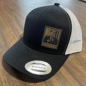 May include: Black and white trucker hat with a brown patch featuring a portrait of a man with the text "You Brissed" in white. The hat has a white mesh back and a black front with a silver label that reads "The Authentic Yupoong Snapback Yupoong Since 1974."
