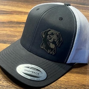 May include: Black and white trucker hat with a black labrador retriever silhouette on the front. The hat has a black front panel and a white mesh back panel. The hat has a black adjustable strap with a silver buckle and the text "The Authentic Snapback Turdoog Since 1974".