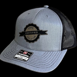 May include: A gray and black trucker hat with a gold patch that reads "OSHA Certified Violator". The hat is adjustable and has a Richardson 112 logo on the side.