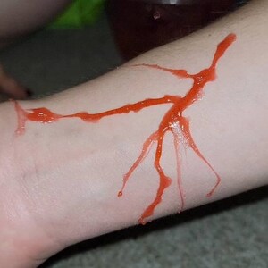 May include: A close-up of a person's arm with a red, fake blood-like substance smeared on it. The substance is in the shape of a jagged line and branches.