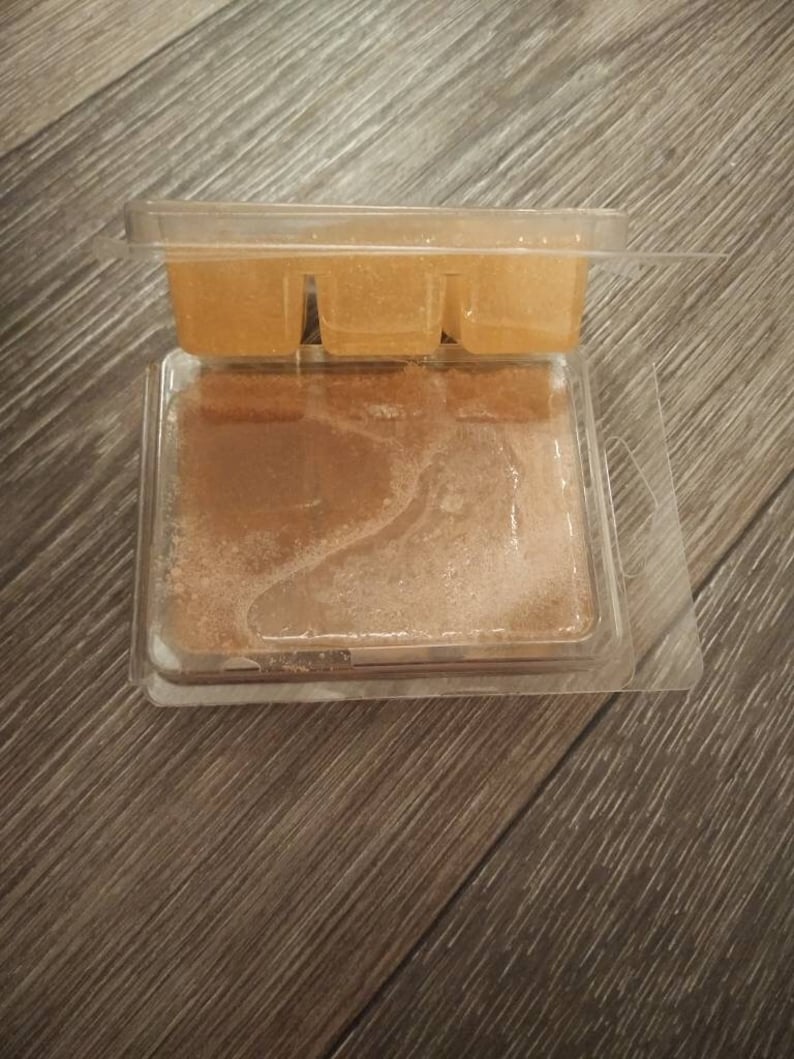 May include: A clear plastic container with three wax melts inside. The wax melts are a light brown color and have a glittery texture.