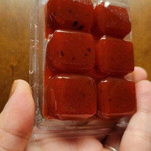May include: A clear plastic container with six red wax melts. The wax melts are square shaped and have a speckled appearance.