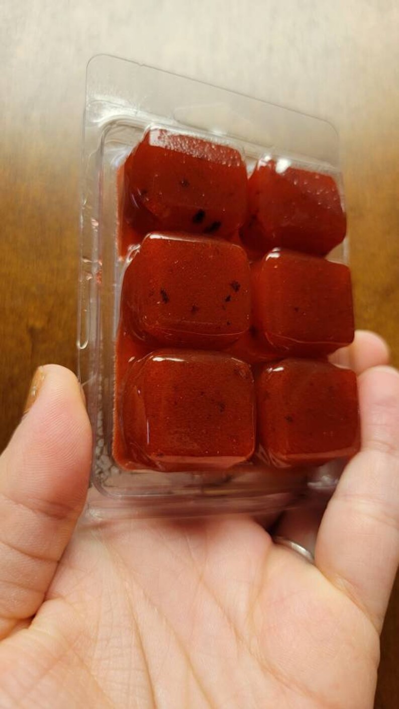 May include: A clear plastic container with six red wax melts. The wax melts are square shaped and have a speckled appearance.