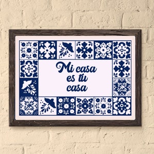 May include: A framed print with a white background and blue and white tile patterns around the edges. The text "Mi casa es tu casa" is written in a blue cursive font in the centre of the print.