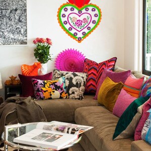 May include: A brown couch with colorful pillows and a large decorative heart with floral embroidery hanging on the wall. The heart is red, orange, green, purple, and white. The couch is decorated with a variety of pillows in different colors and patterns, including pink, red, orange, yellow, green, blue, and black.