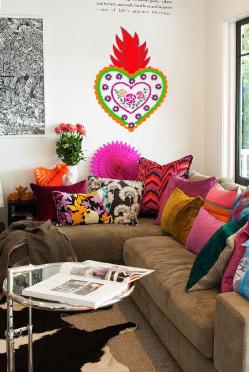 May include: A brown couch with colorful pillows and a large decorative heart with floral embroidery hanging on the wall. The heart is red, orange, green, purple, and white. The couch is decorated with a variety of pillows in different colors and patterns, including pink, red, orange, yellow, green, blue, and black.